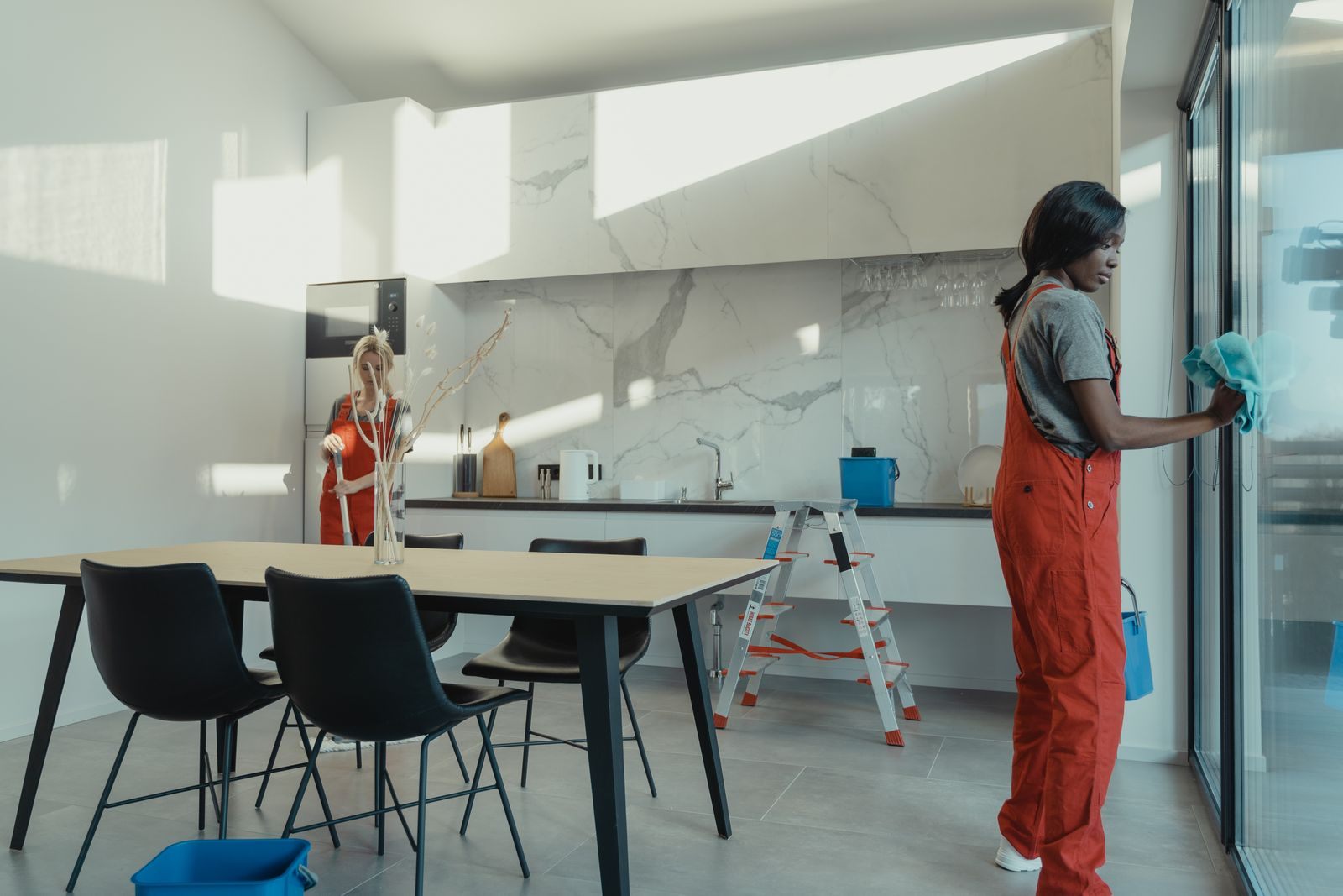 Two women in work clothes cleaning a kitchen with glass doors